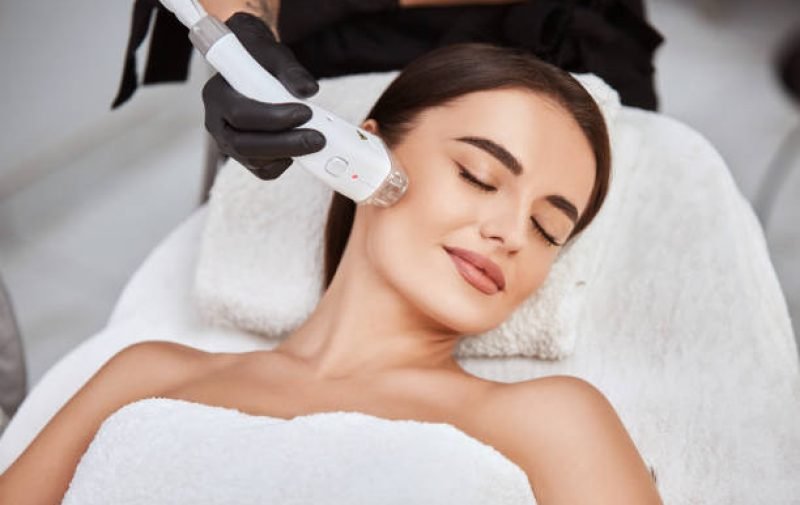 beautiful girl lying in spa salon covering breast with white towel and getting massage for her cheek with professional apparat, lovely woman in beauty and spa concept